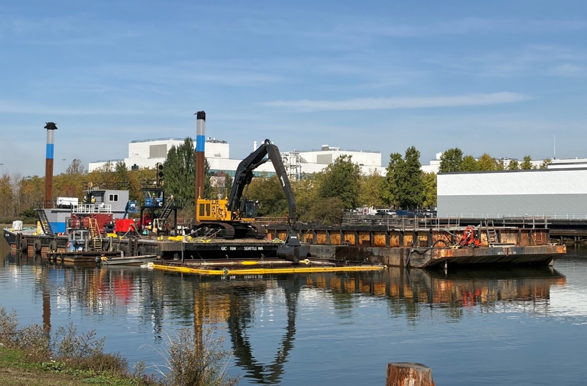2026_1009_SMA 16 dredging_PC King County Heavy equipment, including an excavator, sits on top of a barge on the Lower Duwamish. Next to it rests an empty barge. Trees run the shoreline on the opposite shore and industrial businesses behind that.